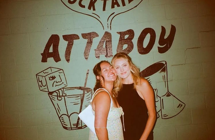 Two friends smiling and posing together in front of a green brick wall with a large cocktail-themed mural and bold lettering, night-out bar vibe.
