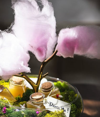 Whimsical party centerpiece with pink cotton candy "trees" perched on a gold branch above moss, tiny corked bottles and a "Drink Me" tag