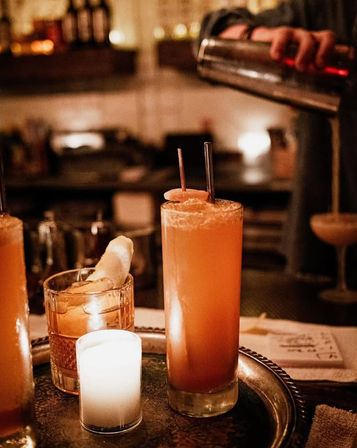 Candlelit cocktail bar scene: a tall sunset‑orange highball with citrus slice and two straws on a tray, a rocks glass with large ice and citrus peel, and a bartender’s hand pouring from a shaker in the blurred background.