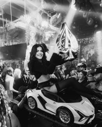 Nightclub party scene: smiling woman in clubwear rides a toy sports car through a crowded dance floor, holding a large spade-shaped bottle under stage lights and cheering guests.