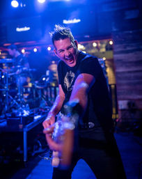 Energetic electric guitarist on a blue-lit stage reaching toward the camera while performing with a live band in a lively bar music venue
