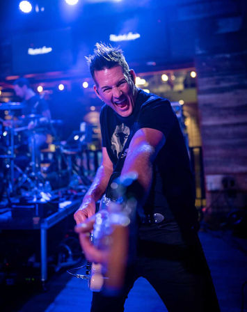 Energetic electric guitarist on a blue-lit stage reaching toward the camera while performing with a live band in a lively bar music venue