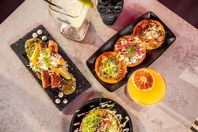 Overhead shot of a colorful Mexican-style spread: three tostadas with shredded slaw and salsa, crispy taquitos with lime and green salsa on a slate, a margarita and an orange cocktail garnished with dried citrus, and a decorative black skull cup.