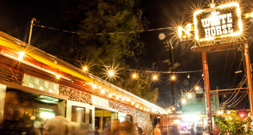 Bustling nighttime outdoor bar patio with string lights, blurred crowd, brick facade and a large glowing marquee sign on a tall pole