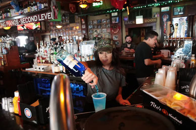 Bartender in a camo cap pouring a vivid blue cocktail into a plastic cup at a lively neighborhood bar with rows of liquor bottles, beer taps, neon signs, and patrons at the counter.