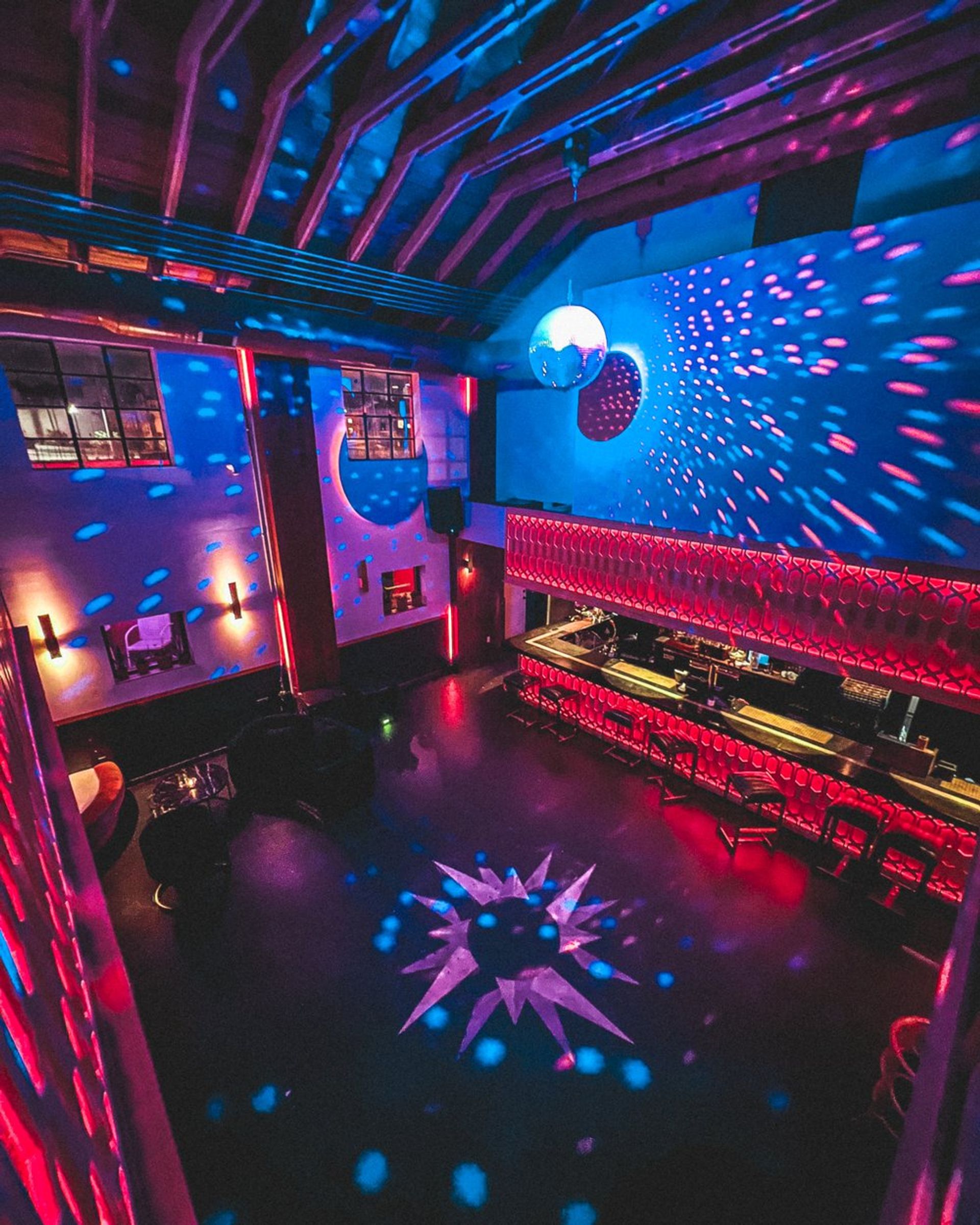 Vibrant two-story nightclub interior with a disco ball casting blue and pink light spots, glowing red bar, and empty dance floor featuring a star-shaped light projection.