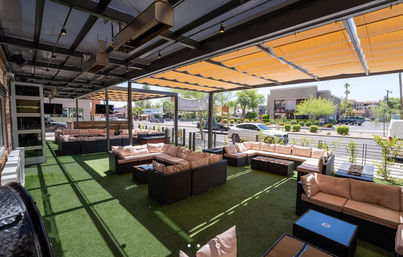 Shaded outdoor patio lounge with dark rattan sectional sofas and beige cushions on green synthetic turf under orange retractable awnings, overlooking a sunny urban street with cars and low-rise shops.
