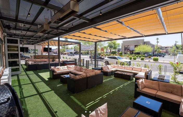 Shaded outdoor patio lounge with dark rattan sectional sofas and beige cushions on green synthetic turf under orange retractable awnings, overlooking a sunny urban street with cars and low-rise shops.