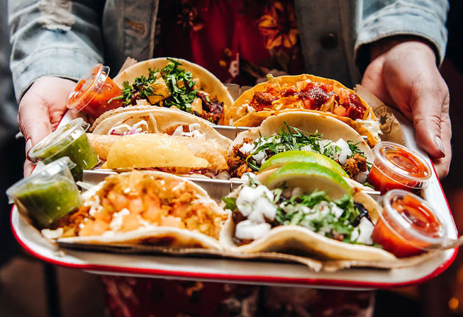Colorful tray of Mexican street-style tacos — assorted fillings with cilantro, diced onion, lime wedges, a fried taco, and green and red salsas in plastic cups.