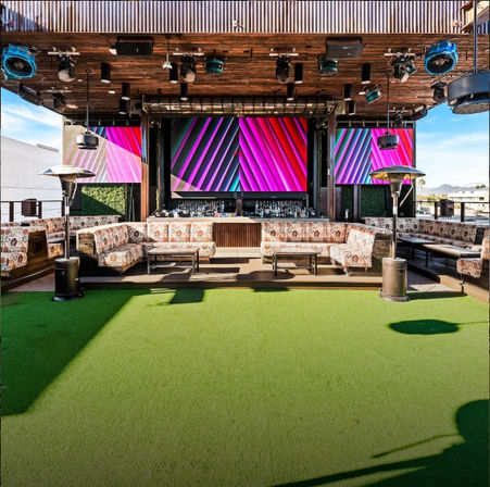 Vibrant urban rooftop lounge with green turf, patterned sectional seating, outdoor heaters and a wooden bar backed by large colorful LED screens.