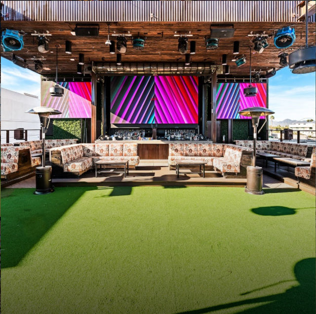 Vibrant urban rooftop lounge with green turf, patterned sectional seating, outdoor heaters and a wooden bar backed by large colorful LED screens.