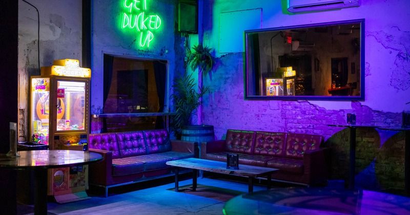 Neon-lit urban lounge with purple and green glow on tufted leather sofas around a low wooden coffee table, a vintage crane arcade machine, exposed brick walls and a large mirror reflecting the cozy bar interior.