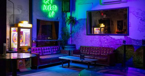 Neon-lit urban lounge with purple and green glow on tufted leather sofas around a low wooden coffee table, a vintage crane arcade machine, exposed brick walls and a large mirror reflecting the cozy bar interior.