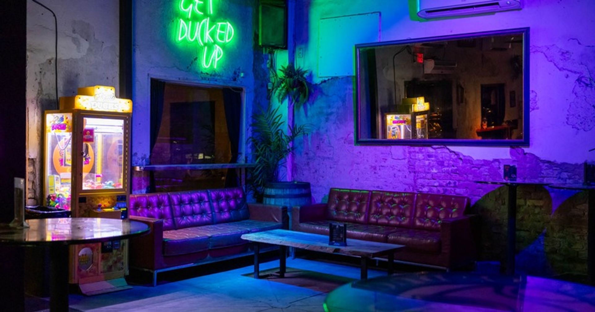 Neon-lit urban lounge with purple and green glow on tufted leather sofas around a low wooden coffee table, a vintage crane arcade machine, exposed brick walls and a large mirror reflecting the cozy bar interior.