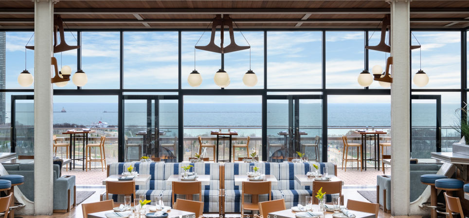 Sunlit coastal restaurant interior with panoramic floor-to-ceiling windows overlooking the ocean, striped blue banquettes, wooden tables set for dining, globe pendant lights, and an outdoor rooftop terrace with high tables and bar stools.