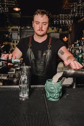 Bartender in a leather apron pouring cocktails into a green tiki mug and a tall ice-filled glass at a dimly lit craft cocktail bar