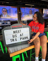 Smiling woman in a red baseball jersey pointing at a marquee sign that reads 'MEET US AT THE PARK' inside a lively sports bar with multiple TV screens and bright green stools.