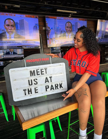 Smiling woman in a red baseball jersey pointing at a marquee sign that reads 'MEET US AT THE PARK' inside a lively sports bar with multiple TV screens and bright green stools.
