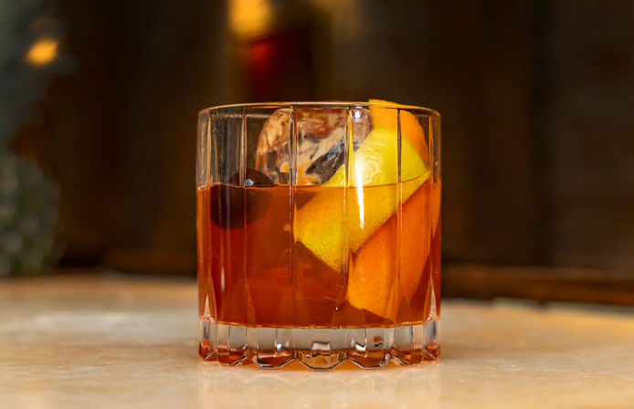 Classic Old Fashioned cocktail in a cut-glass rocks glass with amber whiskey, large ice cube, orange peel and cherry on a marble bar countertop
