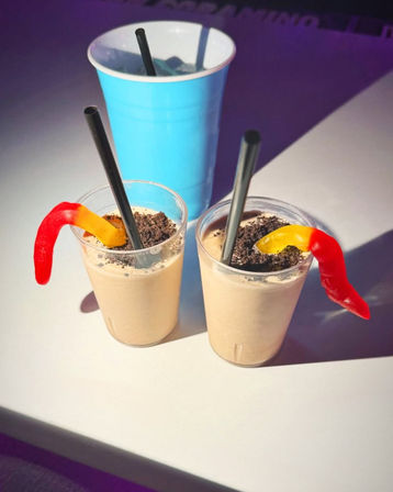 Two creamy milkshake shooters topped with crushed chocolate cookie crumbs and red-and-yellow gummy worms, each with a black straw, beside a tall blue cup on a white party table
