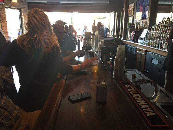 Dimly lit neighborhood bar interior with patrons at a wooden counter, plastic beer cups and a can on the bar, stacked cups and bottles behind the bar, and daylight streaming in from an open front.