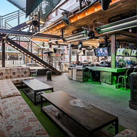 Industrial-chic outdoor bar patio with wooden bar, green accent lighting, multiple TV screens, patterned lounge seating, low coffee tables and metal staircase to an upper level.