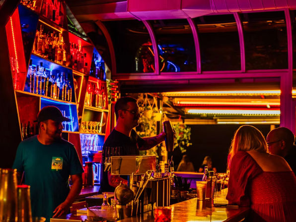 Neon-lit bar interior at night with a bartender shaking a cocktail in front of glowing red and blue bottle shelves, patrons seated at the counter—vibrant nightlife scene.