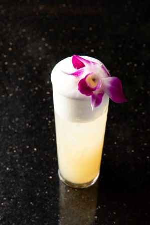Tall tropical cocktail in a highball glass with pale yellow juice, pillowy white foam and a purple orchid garnish, photographed on a speckled black granite countertop.