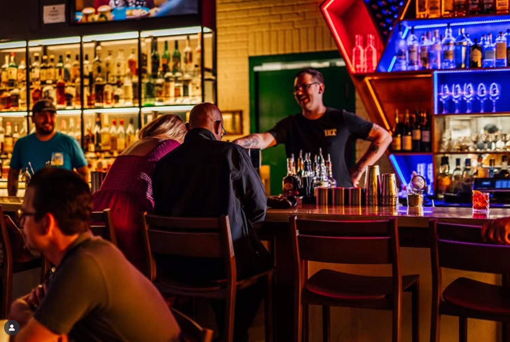 Lively neon-lit cocktail bar with a smiling bartender serving drinks to patrons seated at a wooden counter, colorful backlit liquor shelves and glassware in the background.