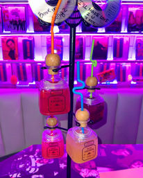 Retro neon bar display of square tequila shot bottles repurposed as colorful cocktails with wooden stoppers and curly straws, purple cassette/CD wall backdrop.