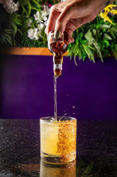 Hand with green nail polish pouring amber spirit into an ice-filled, chili-rimmed yellow cocktail on a black bar counter with a purple backdrop and greenery