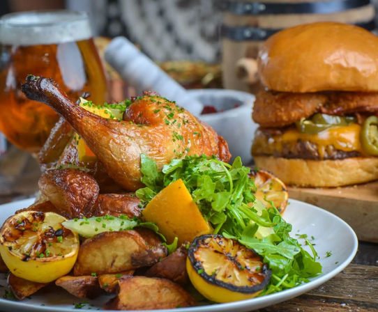 Close-up, mouthwatering pub-style plate with a crispy roasted chicken leg, charred lemon halves, golden potato wedges, peppery arugula, and a cheeseburger with melted cheddar and jalapeños, beer blurred in the background.