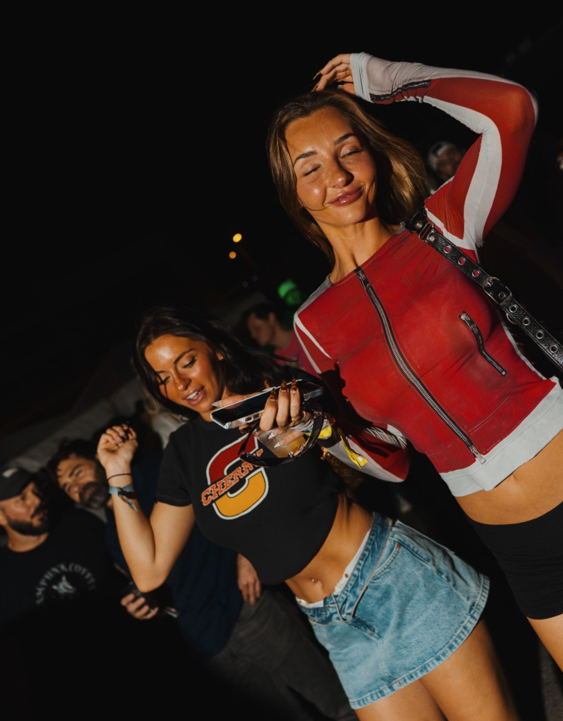 Two young women dancing at a lively outdoor night festival — one in a red zip-up top and black shorts, the other in a black crop tee and denim mini skirt holding a phone and drink, smiling amid a crowded, warm-lit concert vibe.