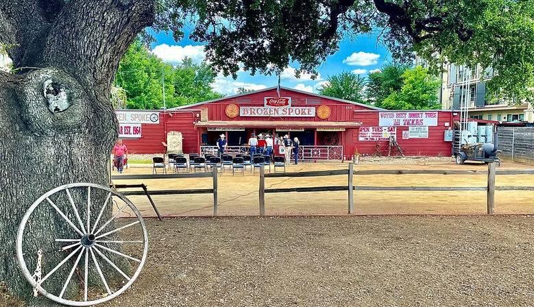 Rustic red Texas honky-tonk and dance hall with open porch, folding chairs in a dirt courtyard, wooden fence, and a large live oak with a vintage wagon wheel in the foreground under a bright blue sky.