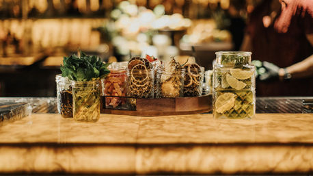 Polished bar counter with mason jars of cocktail garnishes — fresh mint, lime slices, dried citrus and candied fruit on a wooden tray against warm bokeh lights.