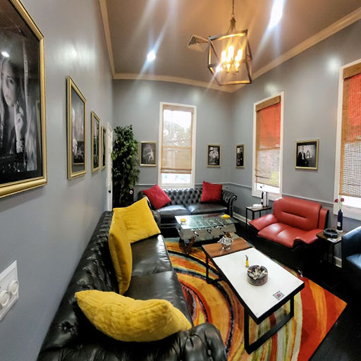 Cozy modern lounge interior with gray walls, tufted black leather sofa with yellow and red throw pillows, red armchair, colorful abstract rug, glass coffee tables, framed black-and-white portraits, potted plant, bamboo blinds and geometric pendant light.