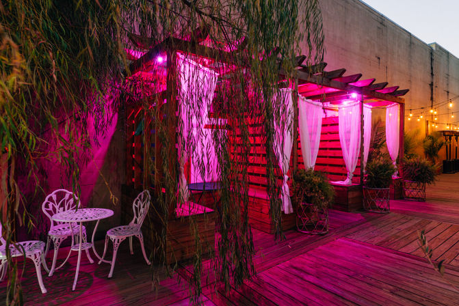 Urban rooftop patio at dusk with wooden cabanas draped in white curtains, pink mood lighting, potted plants, string lights and white wrought-iron table and chairs on a wooden deck