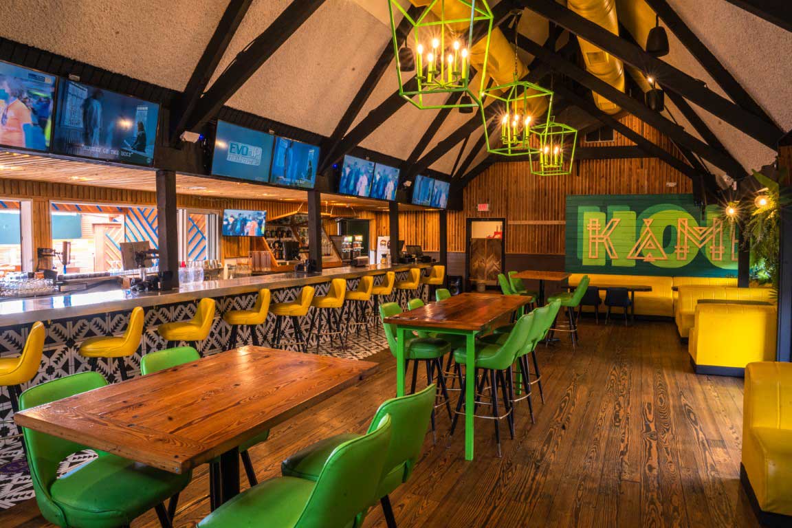 Vibrant bar interior with vaulted beams, wooden floors and tables, green chairs, yellow bar stools, geometric pendant lights, colorful mural and multiple TV screens.