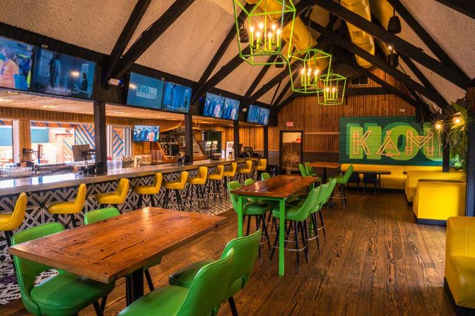 Vibrant bar interior with vaulted beams, wooden floors and tables, green chairs, yellow bar stools, geometric pendant lights, colorful mural and multiple TV screens.