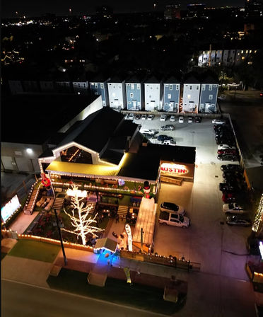 Aerial night view of a festive outdoor dining patio lit with string lights and a glowing white tree, inflatable holiday decorations, adjacent parking lot and a row of townhouse-style buildings under city lights.