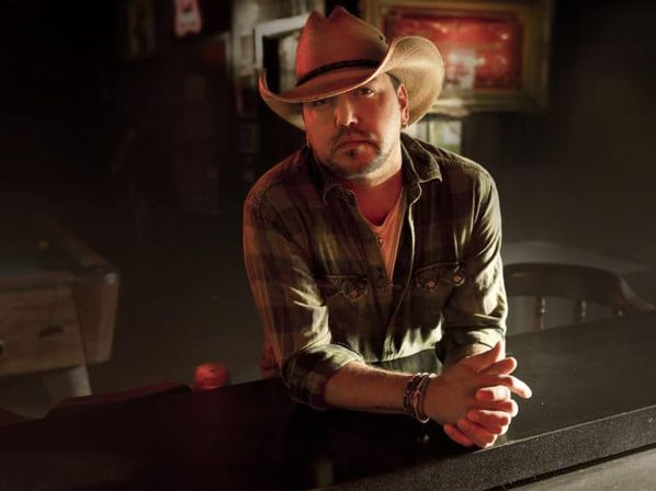 Person in a cowboy hat leaning on a dimly lit rustic country bar counter, wearing a plaid shirt and bracelets with warm, moody lighting