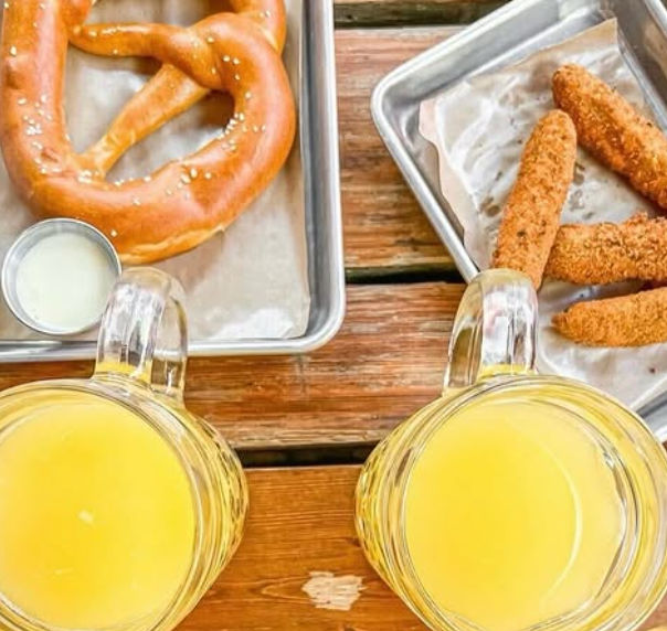 Top-down shot of two cold beer mugs on a wooden patio table with a salted soft pretzel and dipping sauce on one metal tray and crispy breaded mozzarella sticks on another — outdoor beer garden snack spread.
