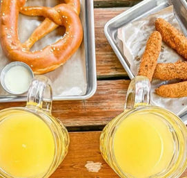 Top-down shot of two cold beer mugs on a wooden patio table with a salted soft pretzel and dipping sauce on one metal tray and crispy breaded mozzarella sticks on another — outdoor beer garden snack spread.