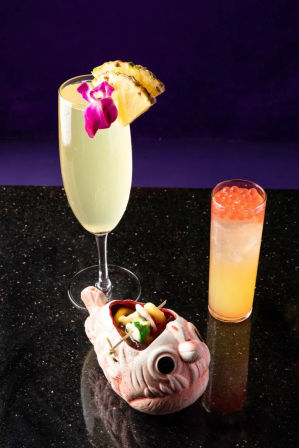 Two tropical cocktails on a black granite bar: a pale champagne flute garnished with pineapple and a purple orchid, and a tall orange highball topped with orange boba, served with seafood skewers in a pink ceramic fish bowl.