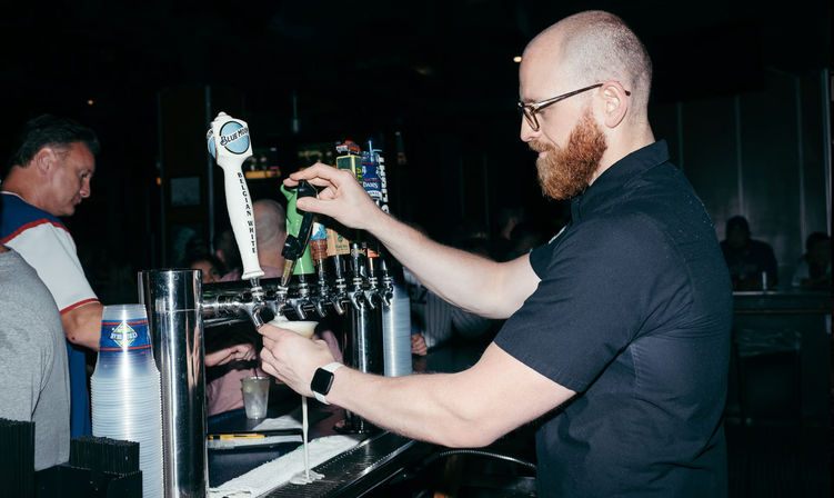 Bearded bartender with glasses mid-pour, filling a plastic cup from a row of draft beer taps at a busy indoor bar.