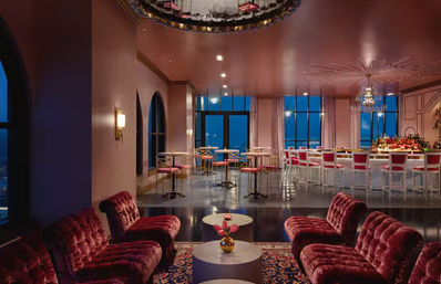 Elevated city-view cocktail lounge at dusk with plush burgundy velvet sofas, round tables, pink-toned ceiling, crystal chandeliers, and a white bar with pink barstools.