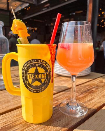 Sunny outdoor patio scene with a tall yellow plastic mug rimmed with chili spice, a red straw and a fruit-and-veggie skewer, next to a stemmed glass of pink-orange cocktail with a strawberry, on a wooden table with string lights in the background.