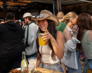 Smiling woman in a straw cowboy hat holding a yellow lime cocktail on a busy outdoor bar patio, wooden table with drinks and snacks in the foreground and string lights and patrons in the background.