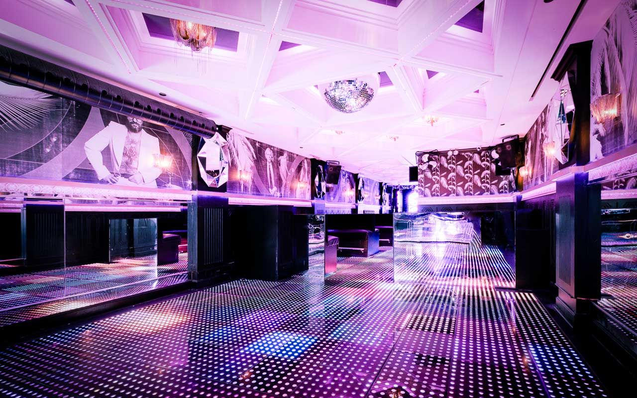 Empty retro-modern nightclub interior with a glowing dotted LED dance floor, mirrored walls, disco ball and pink-purple ambient lighting over plush booths and geometric ceiling.