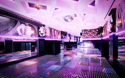 Empty retro-modern nightclub interior with a glowing dotted LED dance floor, mirrored walls, disco ball and pink-purple ambient lighting over plush booths and geometric ceiling.